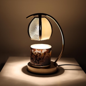 Peut inclure: Un chauffe-cire électrique décoratif avec une monture en métal doré. Il présente une base en céramique à motifs et un abat-jour en verre et crème. La lampe est posée sur une surface blanche.