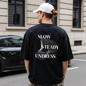 May include: A black short-sleeved t-shirt featuring the white text SLOW STEADY UNDRESS and a white abstract line graphic on the back. The shirt is worn with a white baseball cap, showcasing a casual design with a clean, minimalist aesthetic.