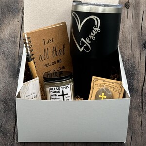 Puede incluir: Una caja de regalo blanca llena de una variedad de artículos, que incluyen un diario marrón con el texto "Let all that you do be done in love", un vaso negro y plateado con la palabra "Jesus" grabada, una vela con el texto "Bless this home with laughter, love, joy", y una pequeña piedra marrón con una cruz dorada.
