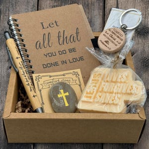May include: A brown gift box filled with a variety of items, including a notebook with the text "Let all that you do be done in love", a pen, a small stone with a cross, a wooden keyring with the text "I can do all things through Christ who strengthens me", and a bar of soap with the text "Jesus forgives saves".