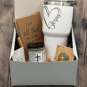 Puede incluir: Una caja de regalo que contiene un vaso blanco con la inscripción "Jesus", un cuaderno de espiral con texto, una vela, un bolígrafo y una piedra de consuelo con una cruz. La caja es gris y está sobre una superficie de madera.