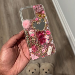 May include: Clear phone case with pink glitter, charms, and a mirror heart. Embellishments include a pink pom-pom, Hello Kitty, a pink bear, a Prada logo, and the name "DELFINA" in gold letters. Other charms include a pink donut, a bottle, and a pink lip.