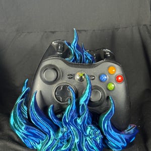 May include: Black Xbox controller with colorful buttons, set in a base of blue and green flames. The flames appear to be 3D printed and add a unique, artistic element to the gaming accessory.