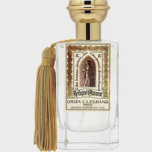 Peut inclure: Un flacon de parfum en verre transparent avec un bouchon doré et un pompon décoratif. L'étiquette porte une illustration et le texte "Relique d'Amour" et "ORIZA L.LEGRAND PARIS MAISON FONDÉE EN 1720". Le flacon est de forme rectangulaire.