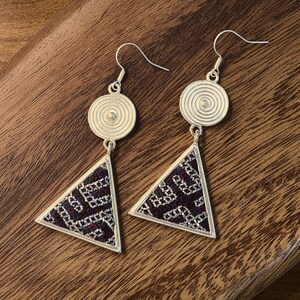 May include: A pair of silver-toned earrings with a geometric design. Each earring features a circular top with a spiral pattern, connected to a triangular pendant with a patterned fabric insert in shades of purple and black. The earrings are displayed on a wooden surface.