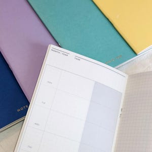 Peut inclure: Un carnet ouvert avec une mise en page d'agenda hebdomadaire, avec des sections pour chaque jour et une liste de tâches. D'autres carnets bleus, violets, turquoise et jaunes l'entourent. Idéal pour la planification.