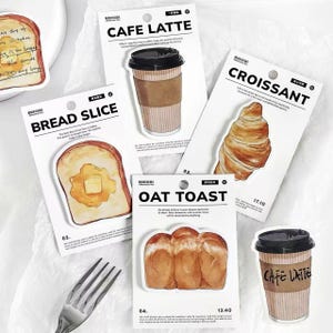 May include: A collection of food-themed stickers featuring a bread slice with butter, a croissant, oat toast, and two coffee cups labeled "Cafe Latte." The stickers are on white cards with black text.