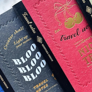May include: Close-up of three travel notebooks with black spines. The notebooks are in shades of gray and pink, with gold foil lettering and embossed designs. The text includes "Travel Notes" and "BLOO BLOO BLOO".
