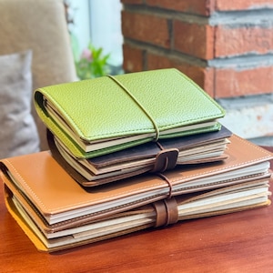 May include: A collection of leather notebooks in shades of green, brown, and tan. Each notebook is secured with an elastic band or a leather strap. The notebooks are stacked on a wooden surface. The notebooks are approximately 10 cm in length.
