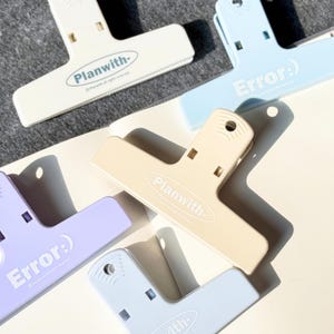 Puede incluir: Un surtido de clips de encuadernación de plástico de colores pastel en tonos crema, azul, lavanda y azul claro. Los clips tienen el texto "Planwith-" o "Error:)" impreso. Los clips están colocados sobre una superficie blanca.