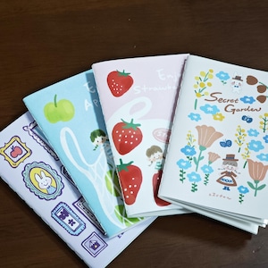 Può includere: Tre quaderni rettangolari con copertine illustrate. Il quaderno a sinistra presenta cornici viola con un coniglio e una mela. Il quaderno centrale è rosa chiaro con fragole rosse e una ragazza. Il quaderno a destra è bianco con fiori e il testo Secret Garden.