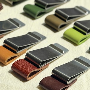 Puede incluir: Una colección de clips para billetes de metal con detalles de cuero en varios colores, incluyendo verde, marrón y rojo. Los clips tienen un acabado de metal cepillado y un diseño curvo.