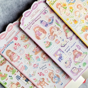 May include: A collection of sticker sheets featuring various illustrations. The stickers depict girls, food, and objects with phrases like "Have a nice day" and "Own the Moment." The color palette includes pink, green, and yellow.