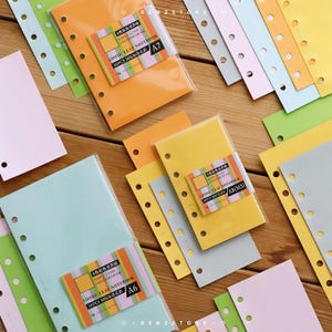 May include: A selection of colourful loose-leaf notebooks in various sizes and shades, including orange, yellow, green, and pink. Each notebook is packaged and labelled with text. The notebooks are arranged on a wooden surface.