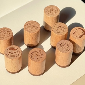 May include: A collection of eight wooden stamps with various food designs. The stamps feature images of a pretzel, bread, a croissant, a slice of cake, a coffee cup, and a loaf of bread. The stamps are cylindrical with a light brown finish.