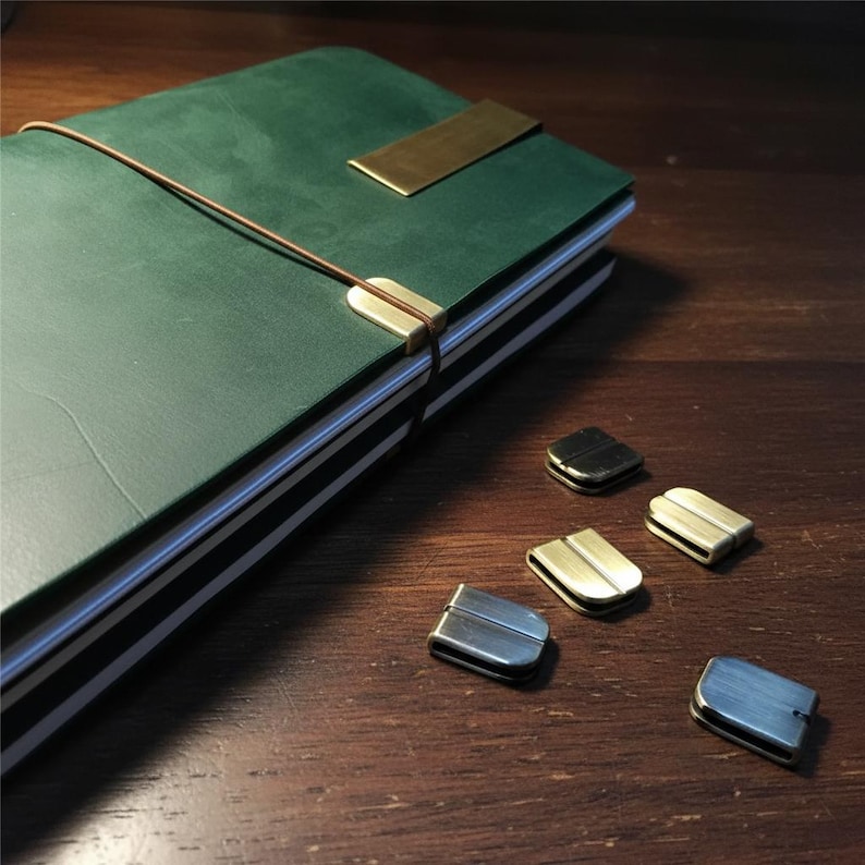 May include: A green leather journal with a brass closure and a brown elastic band. Several metal journal clips in various finishes are scattered nearby. The journal is closed, with the pages visible.