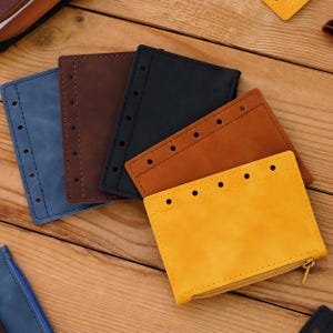 May include: A collection of five rectangular leather pouches in blue, dark brown, black, tan, and yellow. Each pouch features a row of punched holes along one edge for binding and a metal zipper closure along the bottom edge of the yellow pouch.