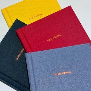 May include: Four hardcover journals in yellow, red, navy, and blue. Each journal has the words "TO DO JOURNAL" embossed in gold on the cover. The journals are arranged in a diagonal pattern, with the yellow journal at the top.
