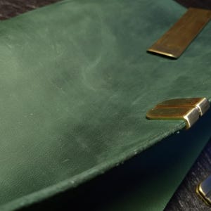May include: Close-up of a dark green leather notebook with brass corner protectors and a brass closure. The leather has a textured appearance. The notebook is open, revealing the inside pages.