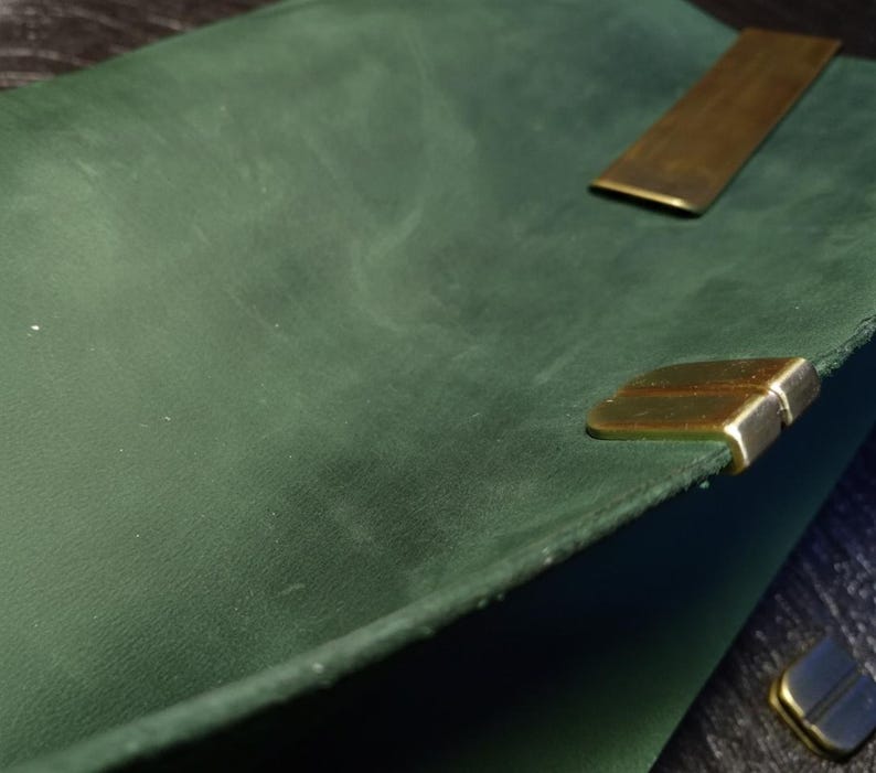 May include: Close-up of a dark green leather notebook with brass corner protectors and a brass closure. The leather has a textured appearance. The notebook is open, revealing the inside pages.