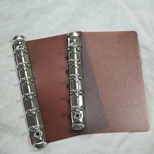 May include: A brown binder cover featuring two vertical silver-toned metal ring mechanisms. The binder has a textured surface and includes six circular rings on each metal spine, designed for holding paper sheets securely in place.