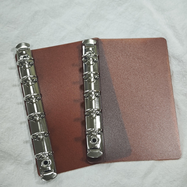 May include: A brown binder cover featuring two vertical silver-toned metal ring mechanisms. The binder has a textured surface and includes six circular rings on each metal spine, designed for holding paper sheets securely in place.