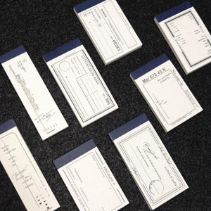 Puede incluir: Una colección de siete blocs de notas rectangulares de papel con encuadernación superior azul oscuro. Cada bloc blanco presenta texto negro único, diseños de estilo vintage y escrituras a mano, incluyendo frases como Letters From Afar.