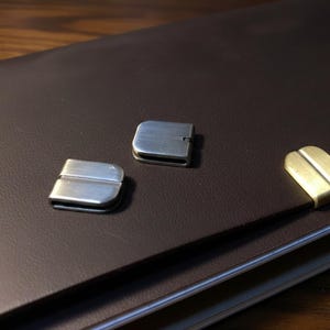 May include: A brown leather portfolio with three metal clasps. Two clasps are silver, and one is gold. The clasps are rectangular with a curved top and bottom. The portfolio is on a wooden surface.