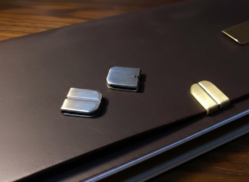 May include: A brown leather portfolio with three metal clasps. Two clasps are silver, and one is gold. The clasps are rectangular with a curved top and bottom. The portfolio is on a wooden surface.