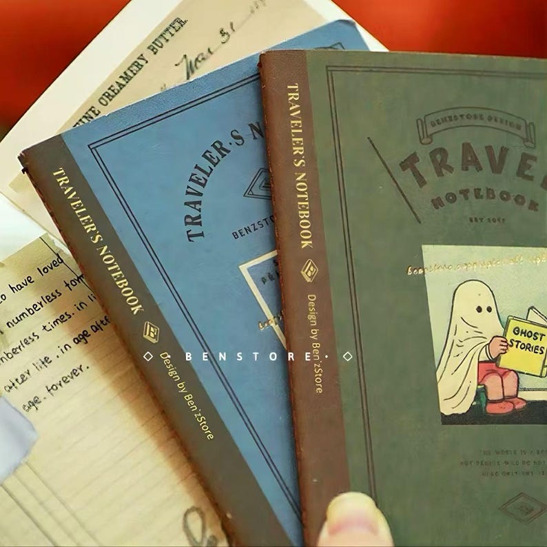 Puede incluir: Varios cuadernos de viaje en azul y verde con lomos marrones. Los cuadernos tienen el texto "Traveler's Notebook" y "Benstore Design" impresos. Uno de los cuadernos presenta una ilustraci&oacute;n de un fantasma con el texto "Ghost Stories".