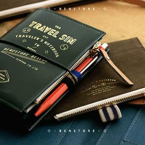 Peut inclure: Carnet de voyage en cuir vert foncé avec lettrage doré « TRAVEL SIM TRAVELER'S NOTEBOOK ». Le carnet est ouvert, révélant un stylo et du papier. Un carnet en cuir marron est à côté. Les carnets sont sur une surface en bois.
