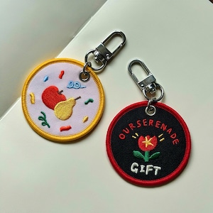 May include: Two circular embroidered keychains with silver metal lobster clasps. One keychain has a light purple background with a red apple, yellow pear, and small colorful shapes. The other is black with a red flower and the text OURSERENADE GIFT.