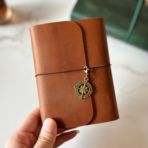May include: A brown leather journal with a compass charm attached. The journal is closed with a brown elastic band. The cover is a solid, warm brown color. The compass charm is a bronze color.