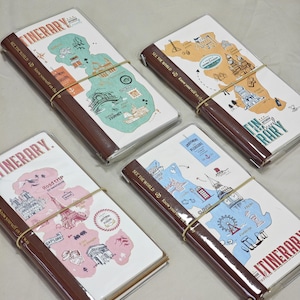 Peut inclure: Quatre carnets de voyage rectangulaires avec des couvertures en similicuir marron foncé et des élastiques. Chaque carnet présente une carte illustrée unique sur la couverture dans des tons pastel, incluant vert, jaune, rose et bleu, avec du texte.