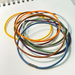 Könnte beinhalten: Eine Sammlung bunter elastischer Haargummis in verschiedenen Farbtönen, darunter Gelb, Orange, Braun, Lila, Grün, Blau und Silber. Jeder Haargummi hat eine runde Form mit einem Metallverschluss. Die Haargummis sind auf einer weißen Oberfläche angeordnet.