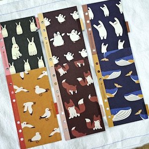 May include: Six rectangular planner inserts with punched holes along the left side. The designs feature repeating patterns of white rabbits on black, seagulls on mustard yellow, penguins on brown, foxes on brown, polar bears on navy, and whales on navy.