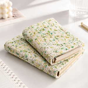 May include: Two rectangular notebooks featuring a textured, woven fabric cover in white with flecks of green, yellow, and blue thread. The notebooks have a ring-bound spine mechanism visible on the side, with cream-colored paper pages inside.
