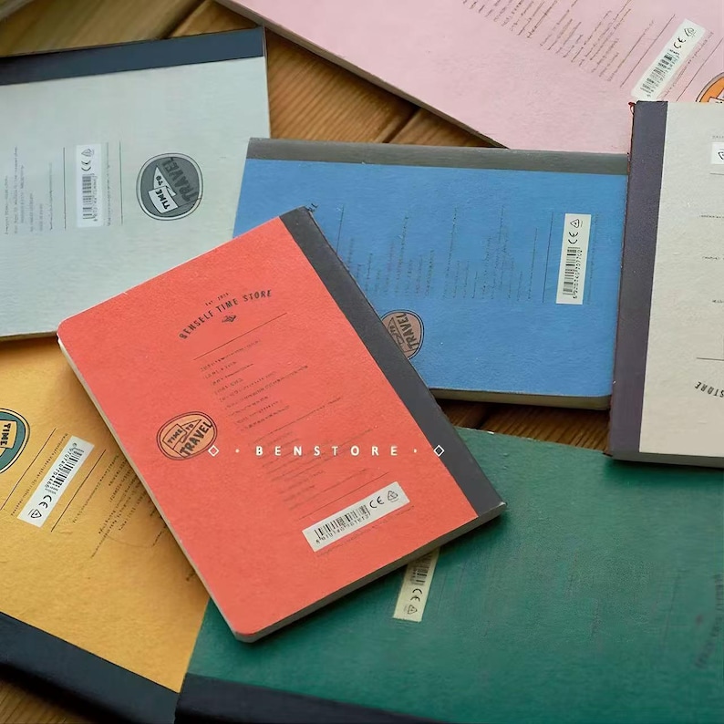 Puede incluir: Una colecci&oacute;n de cuadernos coloridos con lomos negros. Los cuadernos tienen una est&eacute;tica vintage, con las palabras "TIME TRAVEL" y "Benseli Time Store" impresas en las portadas. Los cuadernos son de varios colores, incluyendo rojo, azul, verde y amarillo.