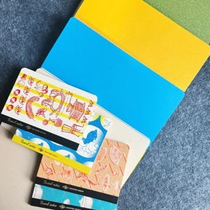 Może przedstawiać: Kolekcja notatników o tematyce podróżniczej z kolorowymi ilustracjami i napisem "Travel notes". Notatniki są ułożone w stos, z żółtym, niebieskim i zielonym tłem. Ilustracje przedstawiają różne obiekty i wzory.