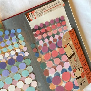 May include: A collection of colorful round stickers in various sizes and shades, including pink, purple, blue, and green. The stickers are displayed in a clear plastic sheet and scattered on a gray surface. The packaging reads "ROUND STICKER".