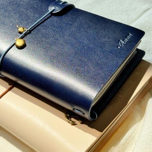 Op de afbeelding: Een close-up van een donkerblauw leren notitieboek met de naam "Anna" in reliëf op de kaft. Het notitieboek is gesloten met een touwtje en een goudkleurige bedel. De tekst "Best Mom est. 96" is zichtbaar eronder.