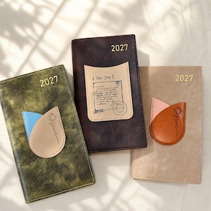 May include: Three rectangular planners for 2027. The left is olive green with a blue and beige petal-shaped pocket featuring a rose drawing. The center is dark brown with a beige pocket showing a letter design. The right is beige with a rose-printed pocket.