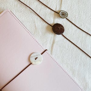 May include: A light pink notebook with a button closure. The notebook is closed with a brown cord and a white button. Two additional buttons, one brown and one silver, are visible above the notebook on a white background.