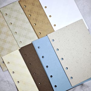 May include: A collection of rectangular paper sheets for a binder, featuring various textures and colors including white, light blue, brown, and cream with grid or speckled patterns. Each sheet has a vertical row of circular punched holes.