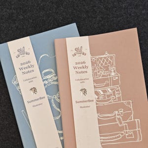 May include: Two 2026 weekly notes planners, one in dusty blue and one in muted pink, each featuring a white paper belly band with the text 2026 Weekly Notes, Collaboration with SummerBee Illustrator, and a white line art illustration of stacked suitcases.