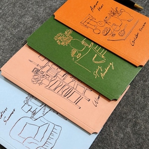 May include: Four colorful notebooks with line art illustrations and handwritten text. The notebooks are orange, green, peach, and light blue. The orange notebook has the text "Autumn Hues" and "Golden Season". The green notebook has the text "Spring Awakening".
