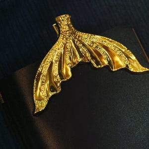 May include: A gold-colored mermaid tail-shaped bookmark. The bookmark is made of metal and has a detailed, textured design. It is resting on a black surface, possibly a book or journal, with a dark blue background.