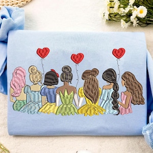 Pode incluir: Uma camisa de frio azul claro com um design bordado de sete figuras femininas com cabelo comprido, vestindo vestidos e segurando balões vermelhos em forma de coração. As figuras estão de costas para o observador.