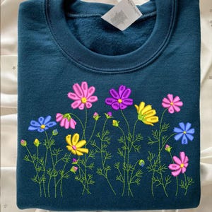 May include: Teal-colored sweatshirt with an embroidered floral design. The embroidery features colorful cosmos flowers in shades of pink, purple, yellow, and blue, with green stems and leaves. The sweatshirt has a crew neck and long sleeves.