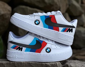Könnte beinhalten: Weiße Turnschuhe mit schwarzen Swooshes und BMW-Logos. Die Schuhe haben blaue, rote und schwarze Streifen an den Seiten mit "M"- und "BMW"-Branding. Die Sohlen sind weiß mit dem Text "AIR".